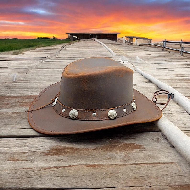 May include: A brown leather cowboy hat with a silver concho band and a brown leather strap. The hat is sitting on a wooden surface.