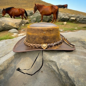 K&ouml;nnte beinhalten: Ein brauner Cowboyhut aus Leder mit einem geflochtenen Band und einer braunen Lederkrempe. Der Hut liegt auf einem gro&szlig;en Felsen mit grasbewachsenem Hintergrund.