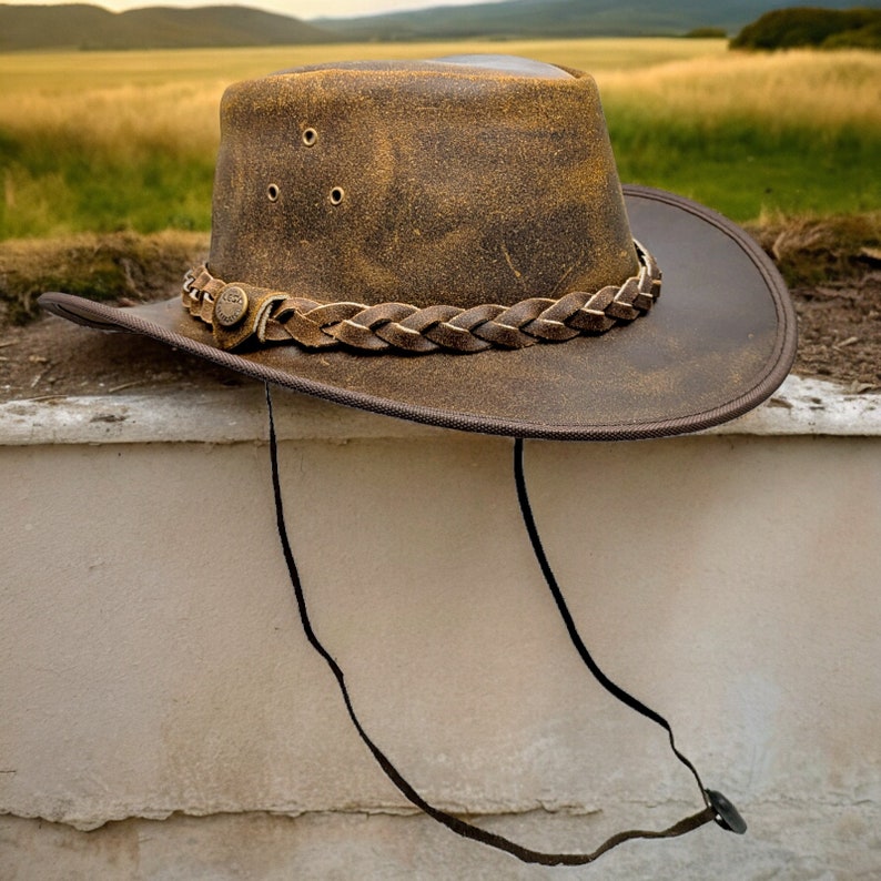 K&ouml;nnte beinhalten: Ein brauner Cowboyhut aus Leder mit einem geflochtenen Lederband und einem braunen Lederkinnriemen. Der Hut hat einen abgenutzten Look und liegt auf einer wei&szlig;en Oberfl&auml;che.