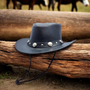 May include: A black leather cowboy hat with a silver concho design and a chin strap. The hat is resting on a wooden log.