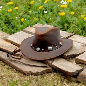 May include: A brown leather cowboy hat with a wide brim and silver concho accents. The hat is sitting on a wooden surface.