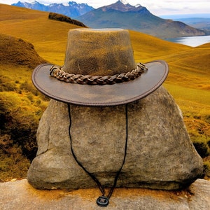 K&ouml;nnte beinhalten: Ein brauner Leder-Cowboyhut mit einem geflochtenen Band sitzt auf einem gro&szlig;en, grauen Felsen. Der Hut ist scharf, w&auml;hrend der Hintergrund eine verschwommene Landschaft aus Bergen und H&uuml;geln ist.