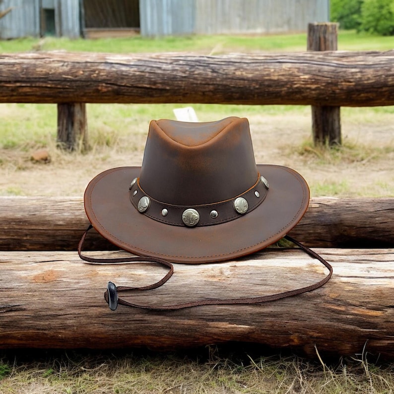 May include: A brown leather cowboy hat with a silver concho band and a brown leather chin strap. The hat is sitting on a wooden fence rail.