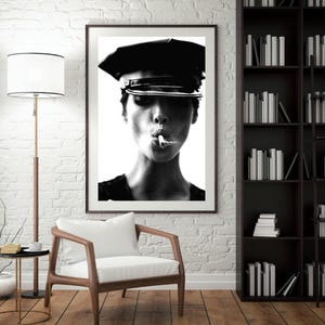 May include: Black and white framed print of a person wearing a black hat and smoking a cigarette. The portrait is a close-up, with the subject's face centered. The image is displayed in a room with a white brick wall, a wooden chair, and a bookshelf.