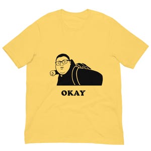 以下が含まれることがあります： メガネをかけた人物の白黒グラフィックと、黒い文字で「OKAY」と書かれた黄色のTシャツ。 グラフィックは、バックパックを背負って横たわっている人物を描いています。