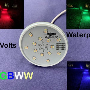 Può includere: Una luce LED rotonda e bianca con 16 LED individuali. La luce è impermeabile e funziona a 24 volt. La luce è etichettata "Rollmop Designs" e ha il testo "RGBWW" stampato sotto. La luce è mostrata mentre illumina un percorso in rosso, verde e blu.