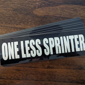 May include: A pile of black rectangular stickers with the text "ONE LESS SPRINTER" in bold white letters. The stickers have a glossy finish and are on a wooden surface.