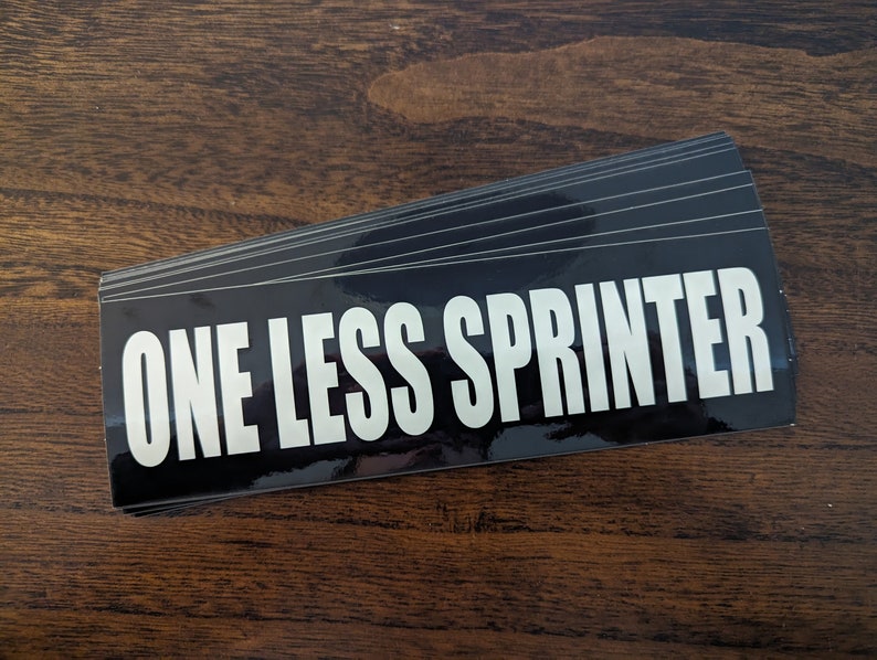 May include: A stack of black rectangular stickers with the text "ONE LESS SPRINTER" in bold white letters. The stickers have a glossy finish and are stacked on a wooden surface.