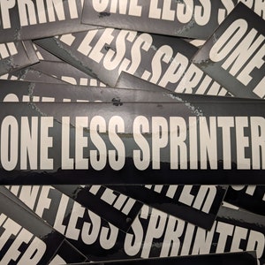 May include: A collection of black rectangular stickers with the text "ONE LESS SPRINTER" in bold white letters. The stickers are glossy and have a slight sheen, with a few showing minor wear and tear.