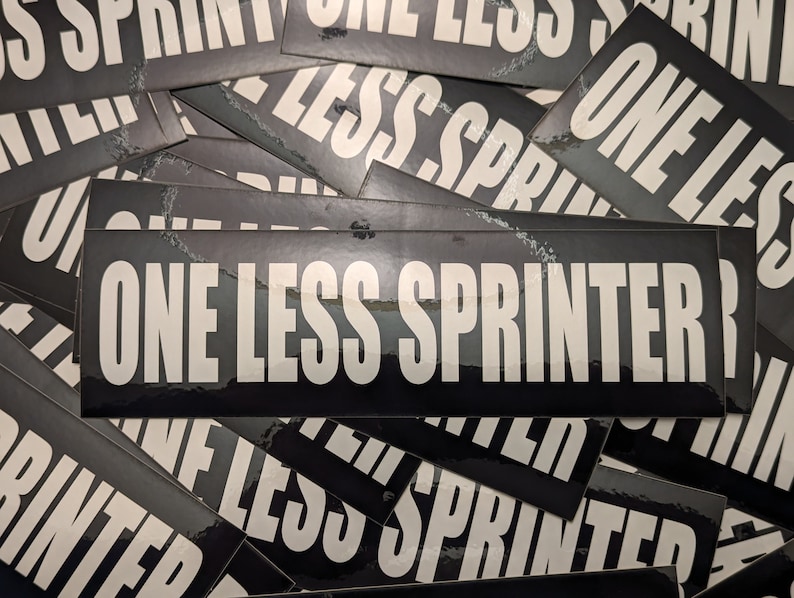 May include: A collection of black rectangular stickers with the text "ONE LESS SPRINTER" in bold white letters. The stickers are glossy and have a slight sheen, with a few showing minor wear and tear.