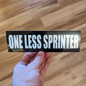 May include: A black rectangular sticker with the text "ONE LESS SPRINTER" in bold white letters. The sticker is held by a hand against a wooden background.