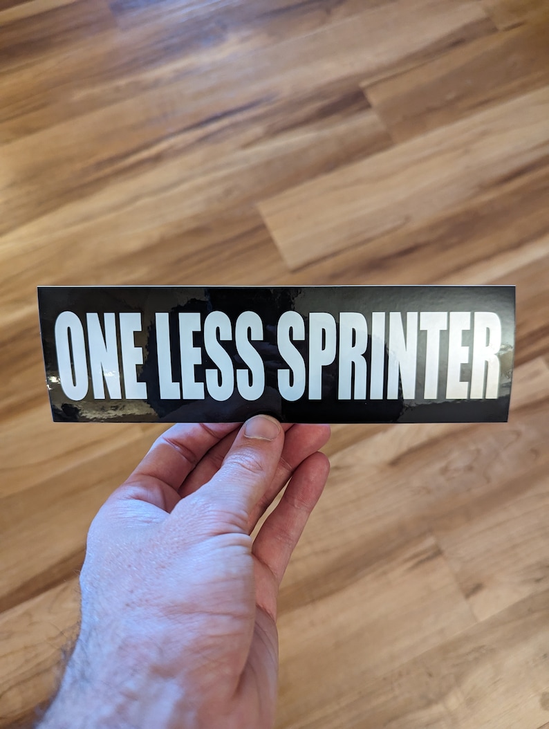 May include: A black rectangular sticker with the text "ONE LESS SPRINTER" in bold white letters. The sticker is held by a hand against a wooden background.
