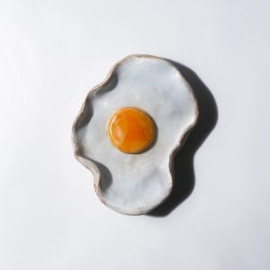 May include: A ceramic plate shaped like a fried egg, with a light blue base and a brown rim. The center features a bright orange yolk. The plate has an organic, wavy edge, and is set against a white background.