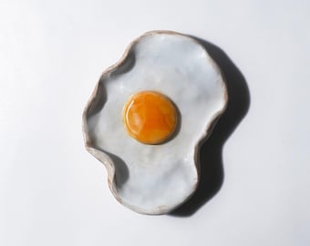 以下が含まれることがあります： 目玉焼きの形をしたセラミックプレート。薄い青色のベースと茶色の縁があります。中央には鮮やかなオレンジ色の黄身があります。プレートは波状の縁があり、白い背景に置かれています。