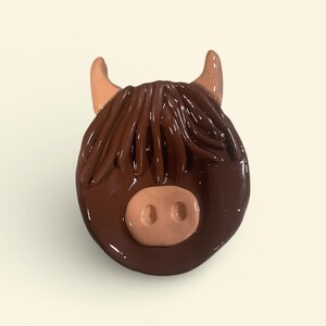 May include: A handcrafted, glossy, brown Highland cow head figurine. The figurine features a light brown nose, tan horns, and long, flowing brown hair. The cow's head is a rounded shape, with a smooth, shiny finish.