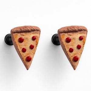 May include: Two pizza slice-shaped wall hooks. Each slice has a brown crust, tan base, and red circular toppings. The hooks are mounted on black hardware against a white background.