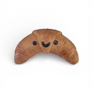 May include: A brown, croissant-shaped object with a smiling face. The face has two black dots for eyes and a curved black line for a mouth. The pastry has a textured surface and is set against a white background.