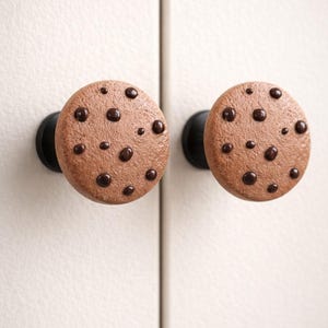 May include: Two round cabinet knobs resembling chocolate chip cookies. The knobs are brown with dark brown chocolate chip details. The knobs are mounted on black hardware against a white surface.