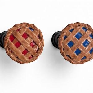 May include: Two decorative cabinet knobs with a pie design. One knob has a red filling, the other has a blue filling. Both knobs have a brown lattice crust and a black base. These knobs add a unique touch to any cabinet or drawer.