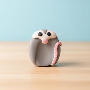 May include: A gray, rounded possum figurine with a pink nose and tail, black eyes, and a white head. The possum is set against a light blue background and rests on a wooden surface.