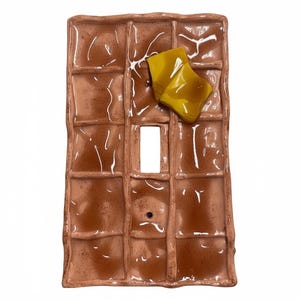 May include: A ceramic light switch cover designed to resemble a waffle, complete with a pat of yellow butter. The brown waffle has a grid pattern and a glossy finish. A fun and unique home decor item.