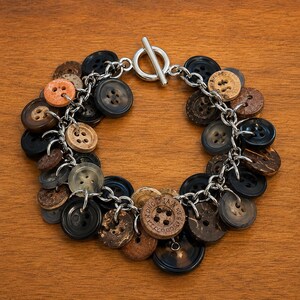 May include: A charm bracelet featuring a silver-toned metal chain with a toggle clasp. The bracelet is adorned with numerous round buttons in various shades of brown, black, and tan, with some buttons displaying visible text and four-hole patterns.