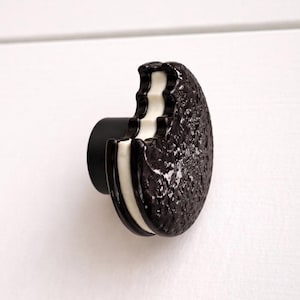 May include: A unique cabinet knob shaped like a chocolate sandwich cookie with a bite taken out. The knob features a black exterior, white filling, and a black mounting post. This decorative hardware adds a playful touch to furniture.