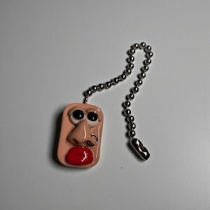 May include: A light pink, rectangular ceiling fan pull chain featuring a cartoon face with black eyes, a nose with a silver nose ring, and red lips. The pull chain is attached to a silver ball chain.