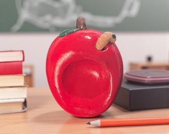 May include: A bright red apple-shaped ceramic bowl with a brown stem and a green leaf. A small, tan worm is emerging from the side. The bowl is on a wooden desk with a pencil and books.