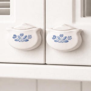 May include: Two white ceramic cabinet knobs with a floral blue design. Each knob has a small, curved handle and a lid. The knobs are mounted on a white cabinet door.
