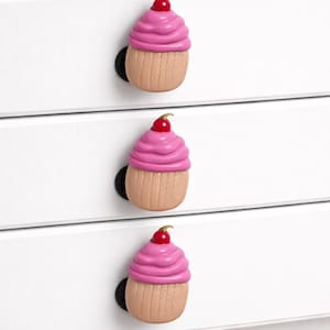 May include: Three cupcake-shaped drawer pulls. Each pull features a pink frosting top with a red cherry and a tan cupcake base. The pulls are mounted on white drawers with black hardware.
