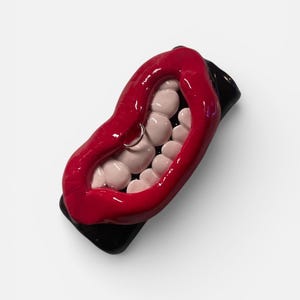May include: A red, glossy, open-mouth sculpture with white teeth and a silver lip ring. The sculpture is mounted on a black base, creating a striking contrast. The overall design is bold and eye-catching, suitable for home decor.