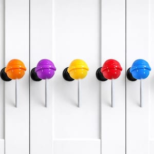 May include: A row of six colorful lollipop-shaped wall hooks. The hooks are in green, orange, purple, yellow, red, and blue. Each hook has a black base and a white stem, mounted on a white surface.