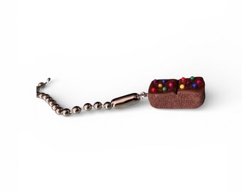May include: A miniature chocolate cake charm with colorful sprinkles on top. The charm is attached to a silver-colored ball chain, perfect for a keychain or zipper pull. The cake is a rich brown color.