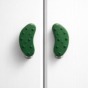 May include: Two green, pickle-shaped cabinet knobs with dark green bumps, mounted on a white cabinet door. The knobs have a shiny finish and are attached to a silver base.