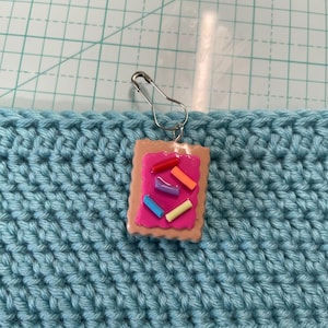 May include: A handmade charm in the shape of a frosted pastry with colorful sprinkles. The charm is attached to a silver safety pin, and is set against a light blue crocheted background.