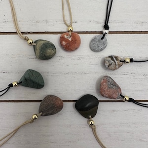 May include: Seven necklaces with different colored stones as pendants. The stones are all smooth and rounded, and they are attached to cords of different colors. The necklaces are all different lengths.
