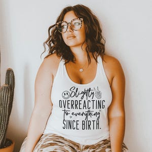 Puede incluir: Camiseta blanca de tirantes con texto negro que dice "Slightly Overreacting To Everything Since Birth." El texto incluye una cara sonriente con rayos y un signo de paz con una mano de esqueleto.