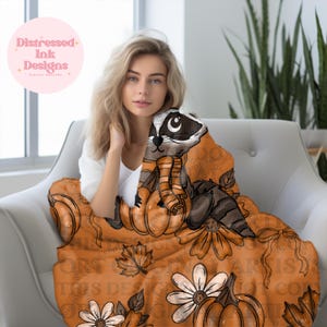 May include: An orange blanket with a raccoon design, pumpkins, flowers, and leaves. The blanket has the text "THIS DESIGN IS NOT FOR COPY". The blanket is draped over a white chair. The blanket is a cozy autumn-themed item.