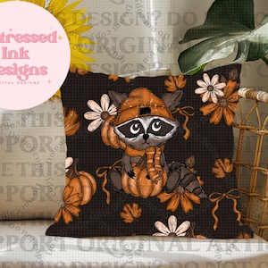 Op de afbeelding: Een decoratief sierkussen met een wasbeer met een oranje hoed en sjaal, die een pompoen vasthoudt. De zwarte achtergrond is versierd met oranje pompoenen, bladeren en bloemen. De tekst "Distressed Ink Designs" staat in een roze cirkel.