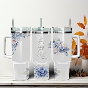 Op de afbeelding: Witte reistumbler van roestvrij staal met een handvat en een doorzichtig deksel. De tumbler heeft een bloemenpatroon met blauwe en roze bloemen en gouden accenten. Op de tumbler staat ook de tekst "She is clothed in strength and dignity and laughs without fear of the future. Proverbs 31:25".