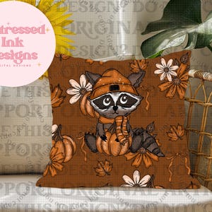 May include: A cushion with a raccoon illustration wearing an orange hat and scarf, holding a pumpkin. The brown pillow has a fall theme with pumpkins, flowers, and leaves. The text "Distressed Ink Designs" is in a pink circle.