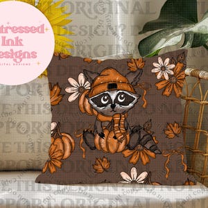 May include: A cushion cover with a raccoon wearing an orange hat and scarf, sitting on a pumpkin. The design has a brown background with autumn illustrations, including pumpkins, leaves, and flowers. The text "Distressed Ink Designs" is in a pink circle.