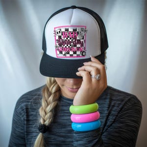 May include: A white and black trucker hat with a pink and black chequered design. The hat has a pink graphic that says "Bad Hair Day Survivor" with a skull, lightning bolt, peace sign, and a pool ball.