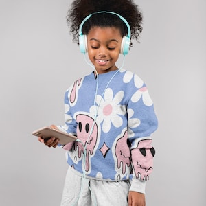 Può includere: Una persona giovane indossa una felpa blu con un motivo a margherita e facce che si sciolgono, pantaloncini grigi e cuffie blu chiaro. Tengono in mano un tablet. Lo sfondo è grigio.