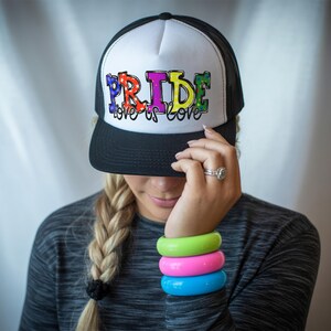 Puede incluir: Una gorra de camionero blanca y negra con un gráfico "PRIDE" de colores arcoíris y el texto "love is love".