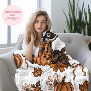 May include: A white blanket with a raccoon wearing an orange scarf and sitting on a pumpkin. The blanket also has orange pumpkins, flowers, and leaves. The text "Distressed Ink Designs" is in a pink circle.