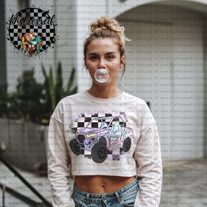 May include: A cream-coloured cropped sweatshirt featuring a colourful graphic of an off-road vehicle with the text "Shop More Designs". The design includes a chequered flag pattern and pastel colours.