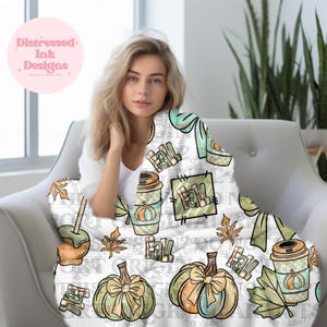 May include: A white blanket with a fall-themed pattern featuring pumpkins, coffee cups, and the word "Fall." The blanket has a grid-like background and is decorated with autumn leaves and bows. The blanket is draped over a chair.