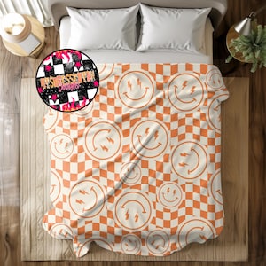 Può includere: Una coperta color crema con un motivo ripetuto di quadrati a scacchiera arancioni e facce sorridenti con accenti di fulmini. La coperta è su un letto con cuscini bianchi.